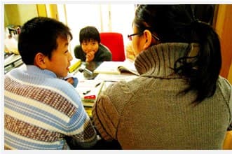 一名戴眼镜的女义工侧身为一个男孩讲题,另一个男孩在桌子另一头看书